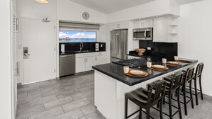 A modern kitchen with white cabinetry, a central island with bar stools, stainless steel appliances, and a view of the ocean through a window. - Ocean View 1-bed/2-bath Penthouse Suite at Waikiki Shore! (Honolulu)