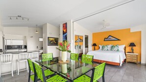 A modern hotel room with a glass dining table, green chairs, a bed with orange bedding, and a kitchen area. - Ocean View Penthouse Suite, Steps to Beach, Sleeps 6! (Honolulu)
