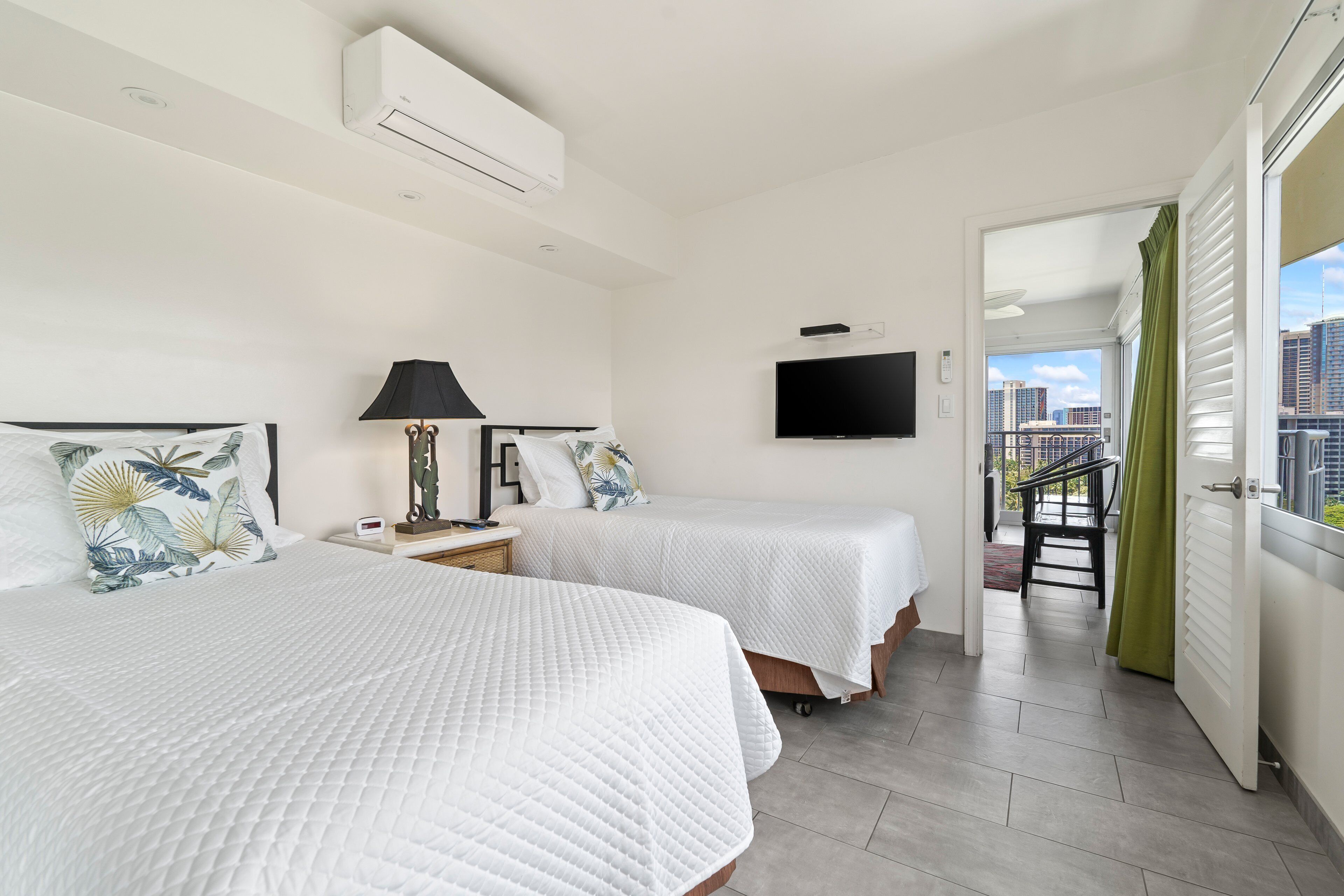 A hotel room with two beds, a TV, and a balcony with a view of the city.