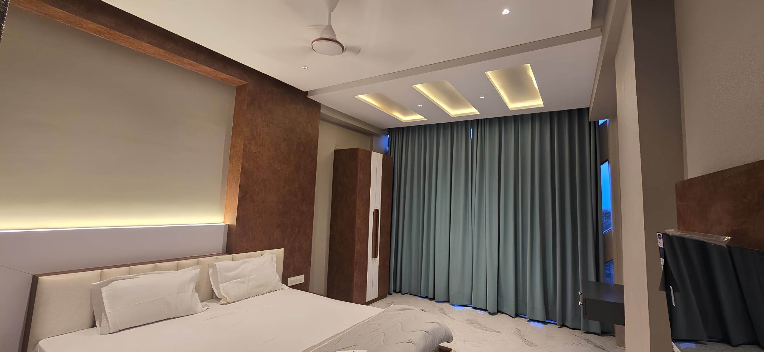 A modern hotel room with a large bed, recessed ceiling lighting, and a view of the city through the window.