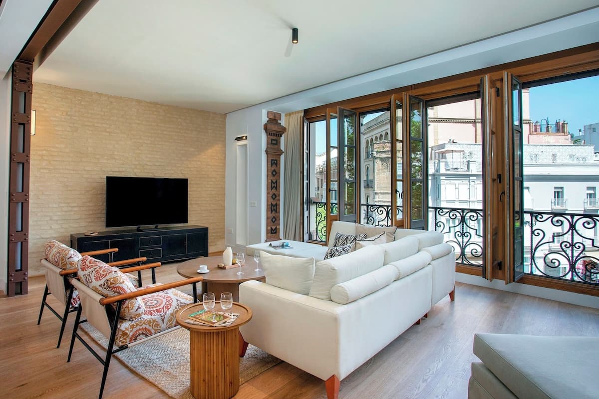 A spacious living room with a large sofa, a television, and a balcony with a view of the city.
