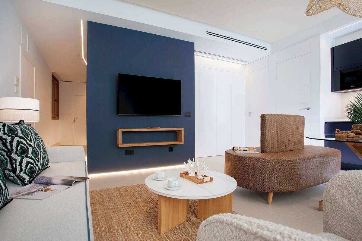 A modern living room with a flat-screen TV mounted on a dark wall, a round wooden coffee table with white cups, a brown woven chair, and a white sofa with patterned cushions.