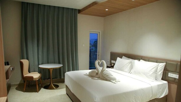A hotel room with a large bed, a small round table, a chair, and a view of the city through a window. - LEAF HOTEL (Tiruvannamalai)