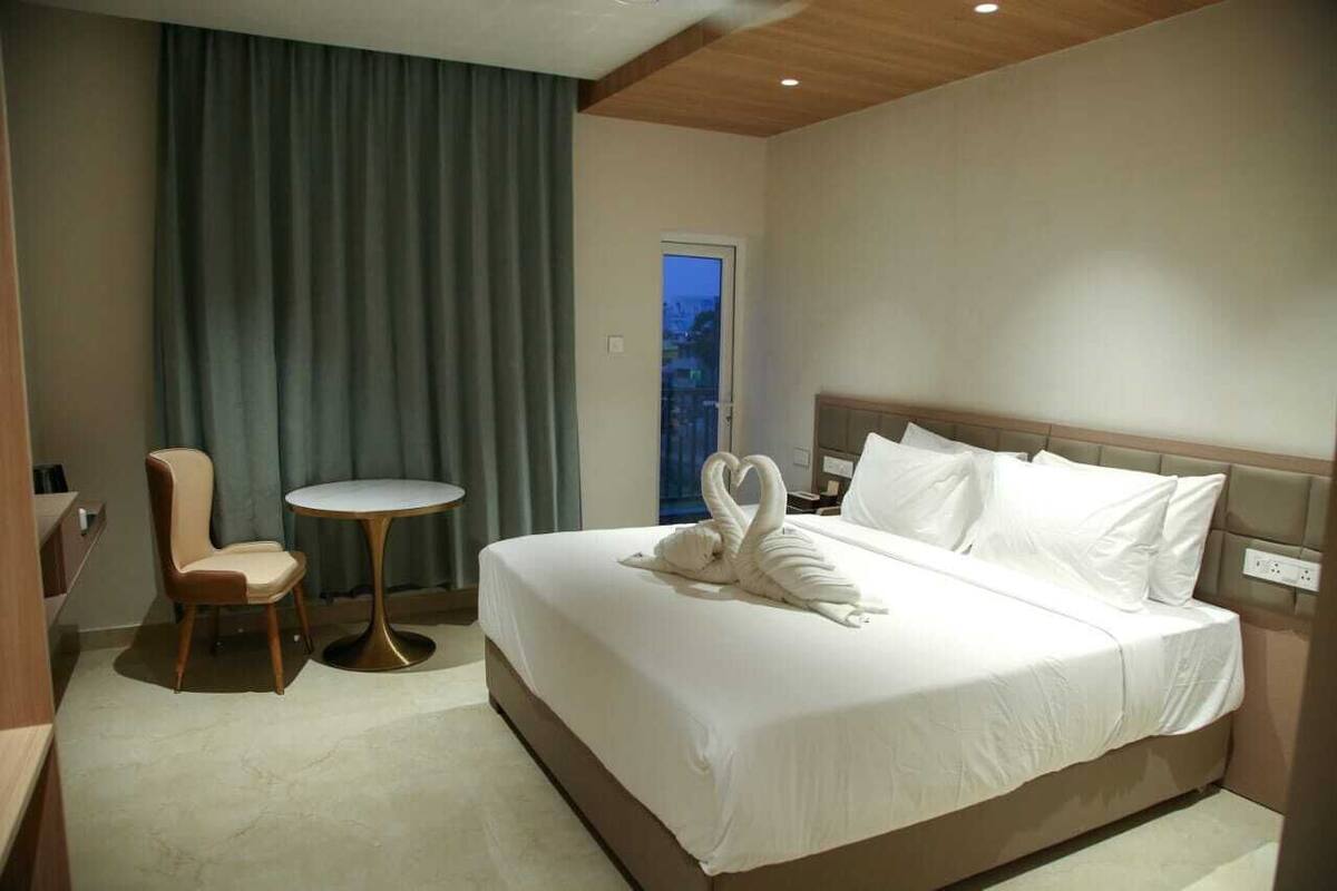 A hotel room with a large bed, a small round table, a chair, and a view of the city through a window.