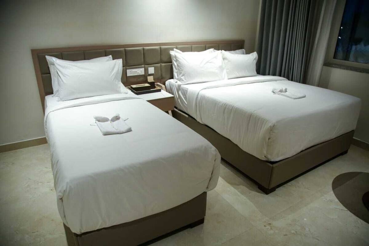 A hotel room with two beds, each with white linens and pillows, a nightstand with a phone and a lamp, and a window with curtains.