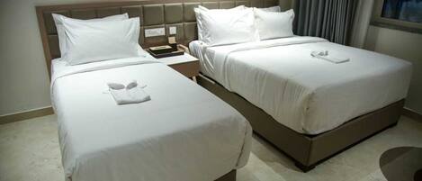 Habitación de hotel con dos camas, cada una con sábanas y almohadas blancas, una mesita de noche con teléfono y lámpara, y una ventana con cortinas.