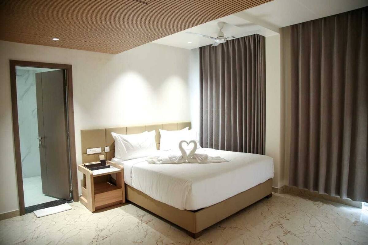 A hotel room with a large bed, a wooden nightstand, a ceiling fan, and a view of the outside through a window with curtains.