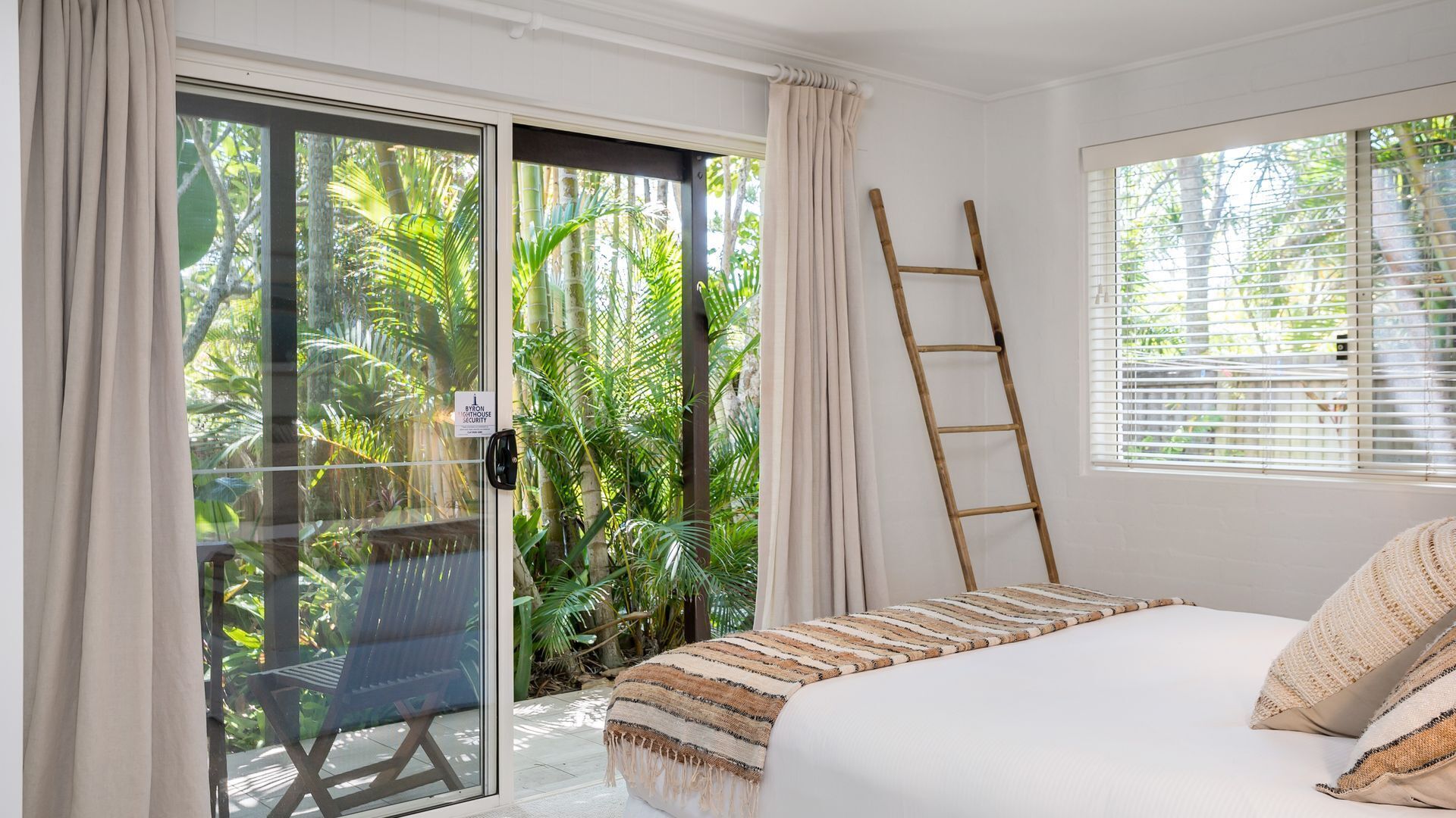 A bedroom with a bed, a window with blinds, a door with a security sign, and a view of tropical foliage.