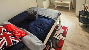 Un dormitorio con cama, mesita de noche, radiador, ventana con cortinas y planta en maceta.