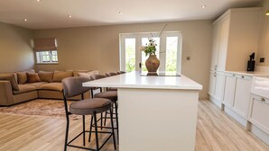 A modern kitchen with a central island, bar stools, a dining area with a sofa, and a view through large windows. - Egerton Cottage (Crewe)