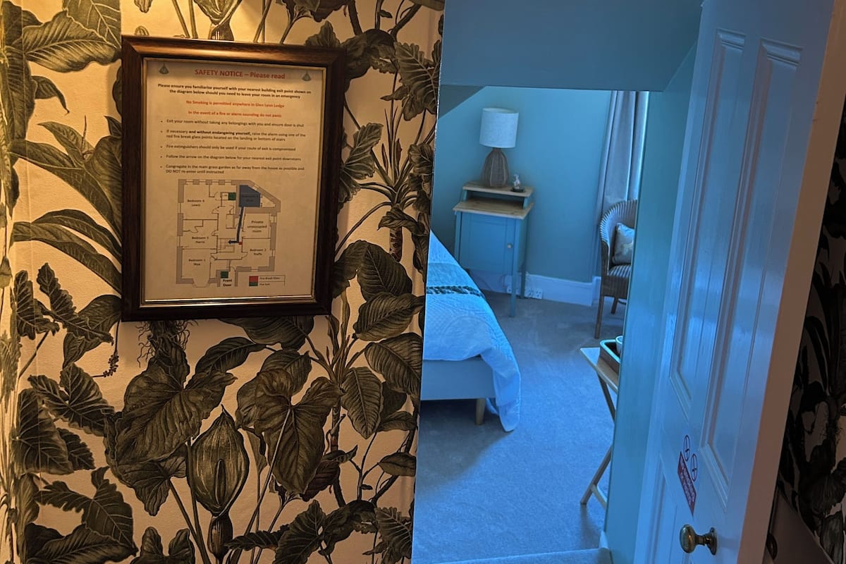 Un couloir orné d’un panneau d’avertissement encadré, un mur décoré d’un motif végétal et une porte donnant sur une chambre à coucher équipée d’un lit et d’une table de chevet.