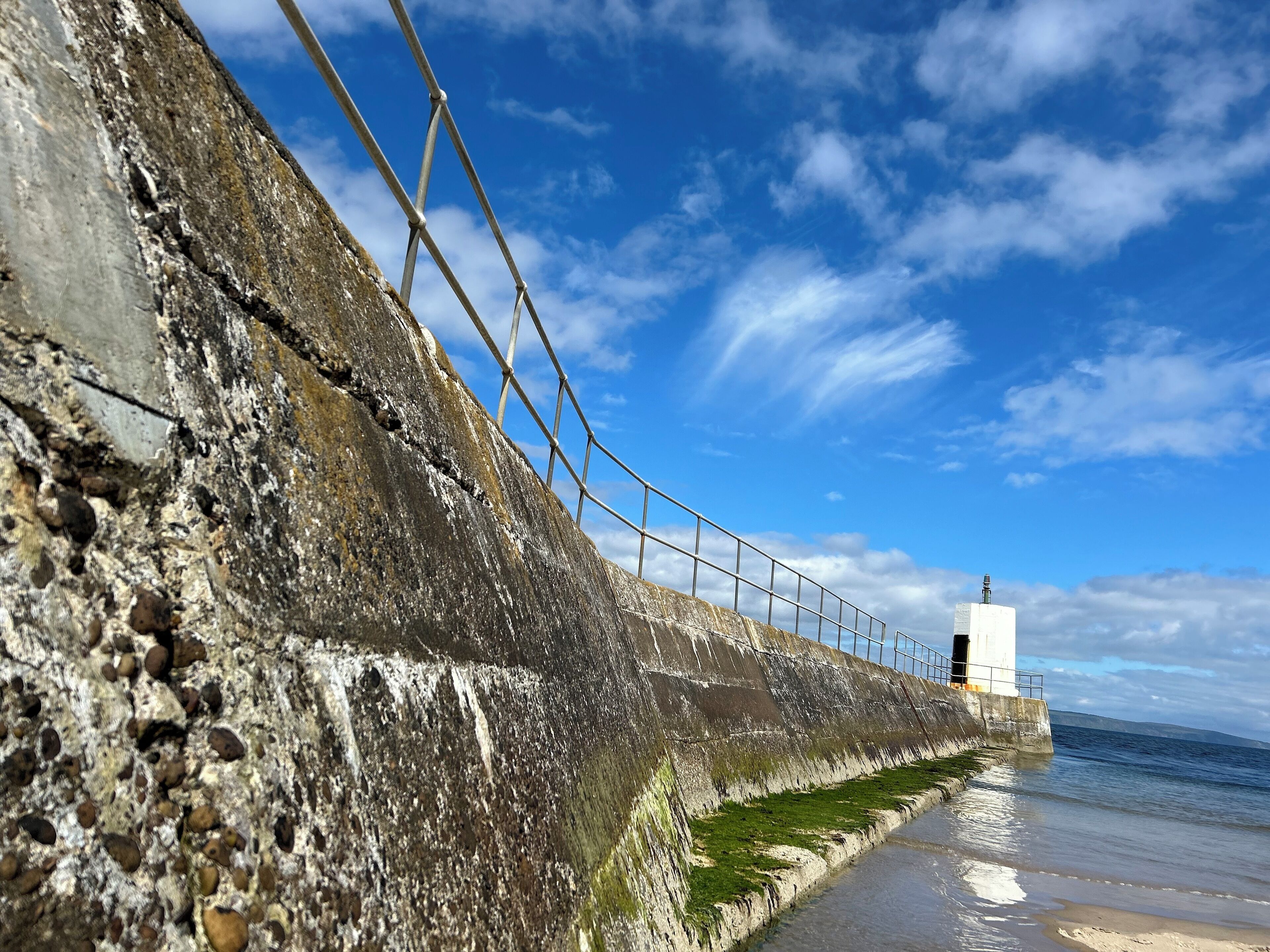 A coastal walkway with a concrete wall, metal railing, and a lighthouse in the distance.