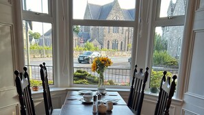 Une table à manger avec des chaises, un vase rempli de fleurs et une vue sur un bâtiment à travers de grandes fenêtres.