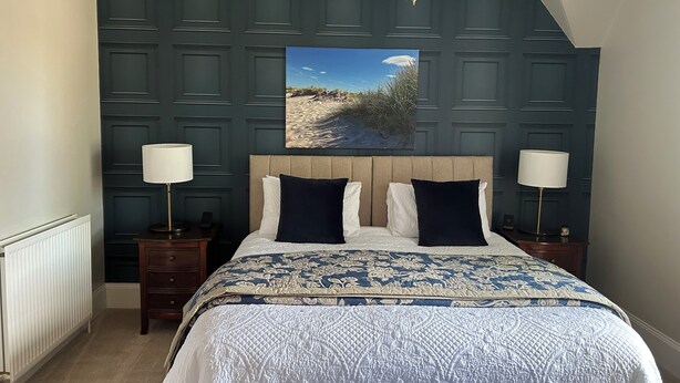 Une chambre à coucher avec un grand lit, deux lampes de chevet, un lustre, une tête de lit ornée d’un tableau et un radiateur.
