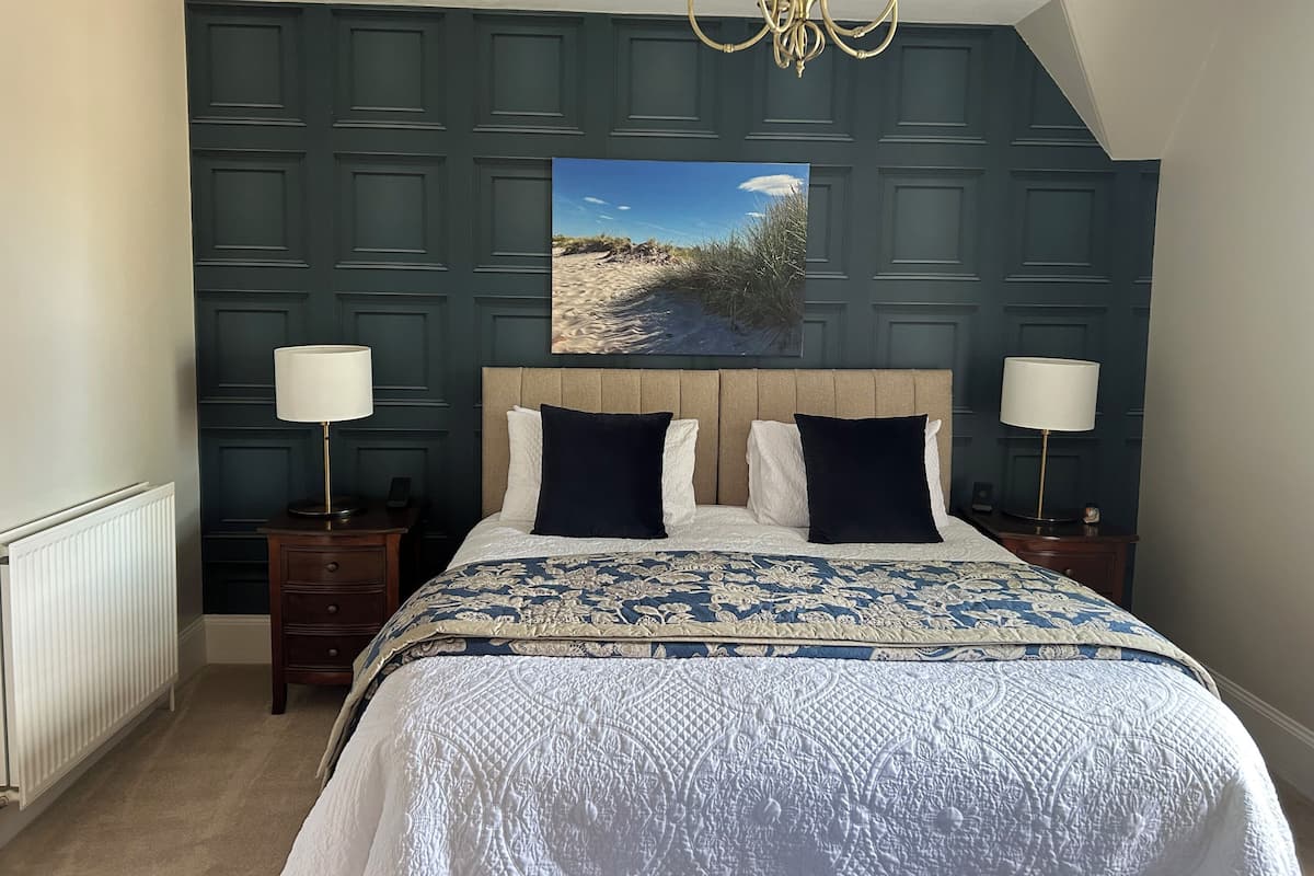 Une chambre à coucher avec un grand lit, deux lampes de chevet, un lustre, une tête de lit ornée d’un tableau et un radiateur.