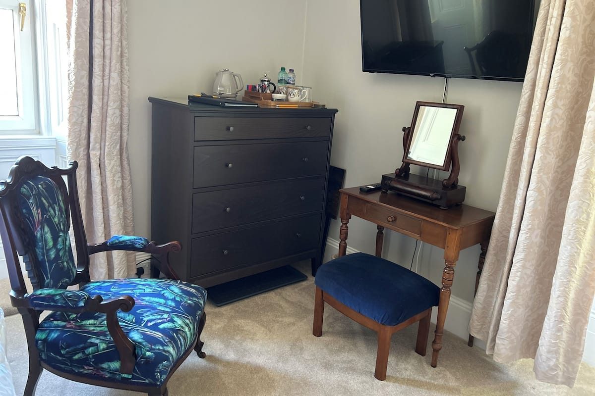 A room with a dark wooden dresser, a desk with a television, a chair with a blue patterned cushion, and a window with curtains.