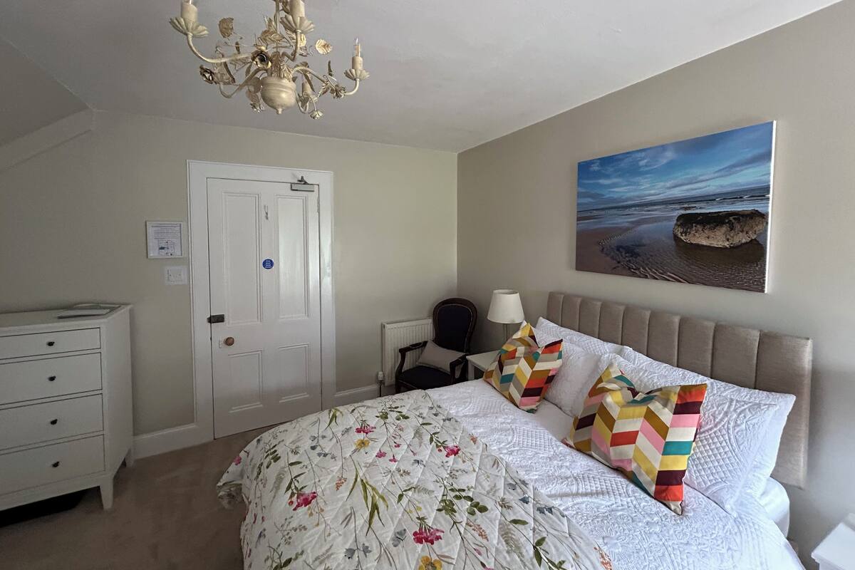 Une chambre à coucher avec un lit, une commode, un lustre, un tableau représentant une plage et une porte.