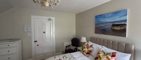 A bedroom with a bed, a dresser, a chandelier, a painting of a beach, and a door.