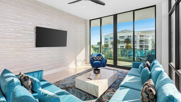 A modern living room with a large window, a ceiling fan, and a flat-screen TV mounted on the wall. - Hi3051hl204: 3 Bed, Ocean Breeze (Melbourne Beach)
