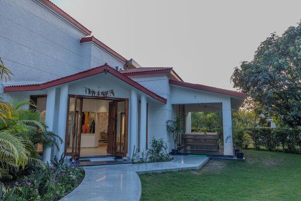 A building with a white exterior and red roof, featuring a covered entrance and a garden area with palm trees and shrubs. - Sukoon A Family Villa (Rishikesh)
