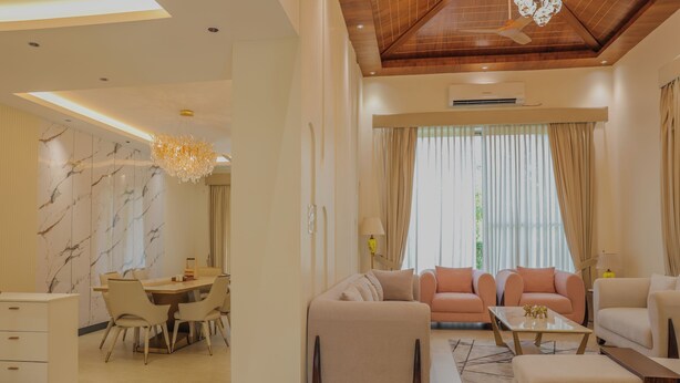 A modern living room with a sofa, ottoman, and coffee table. There is a dining area with a table and chairs. The room has a wooden ceiling and large windows with curtains. - Sukoon A Family Villa (Rishikesh)