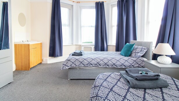 A bedroom with two beds, a bedside table with a lamp, a wooden cabinet, and a large window with curtains. - Thornbury Guest House Room 4 (Southampton)