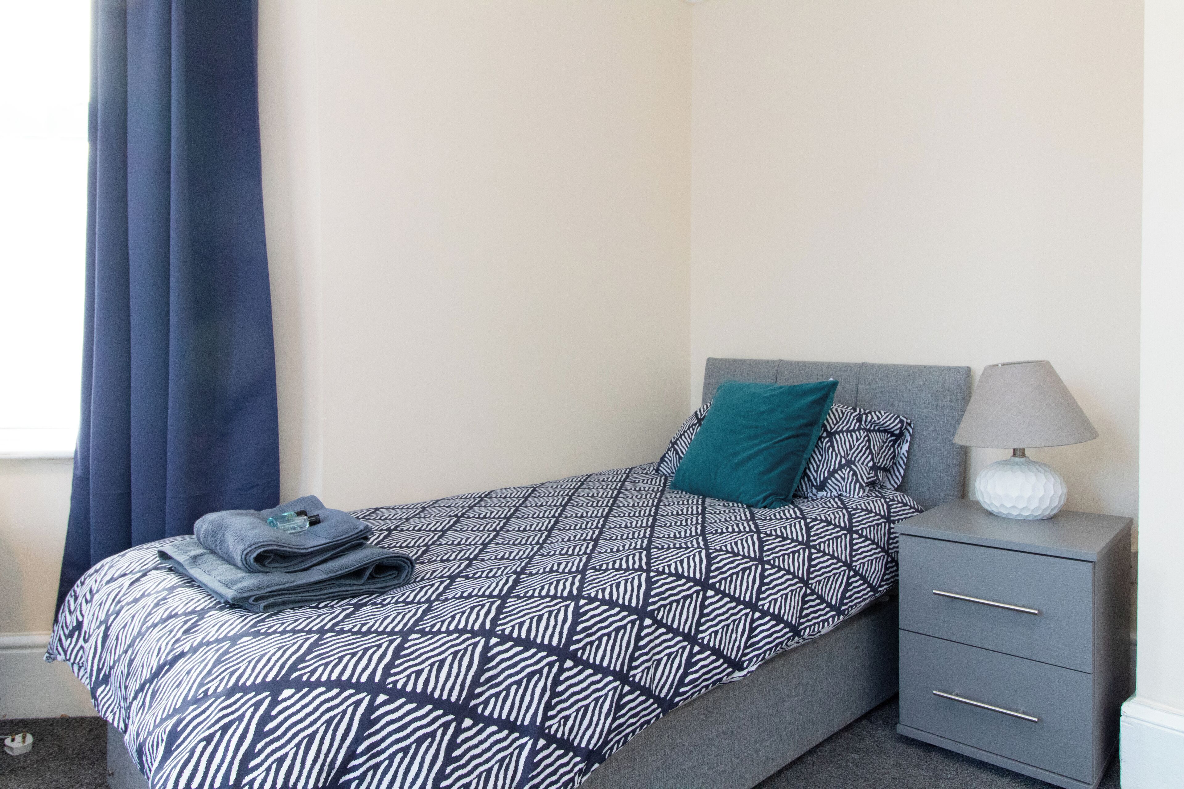 A neatly made bed with a geometric patterned bedspread, a teal pillow, and a folded towel on the side. There is a gray bedside table with a lamp and a decorative item on top.