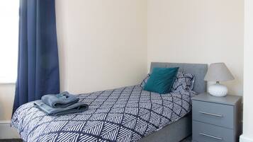 A neatly made bed with a geometric patterned bedspread, a teal pillow, and a folded towel on the side. There is a gray bedside table with a lamp and a decorative item on top.