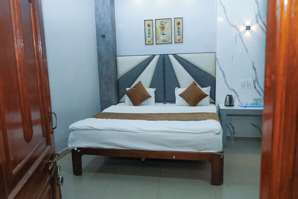A neatly made bed with a headboard, a bedside table with a kettle, and a wall with three framed pictures. - Hotel Pleasure Landmark (Ujjain)