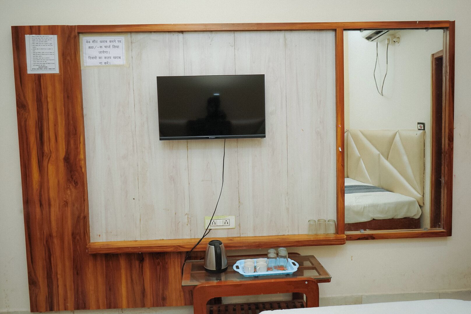 A room with a television mounted on the wall, a bedside table with a kettle and cups, and a bed with a headboard.