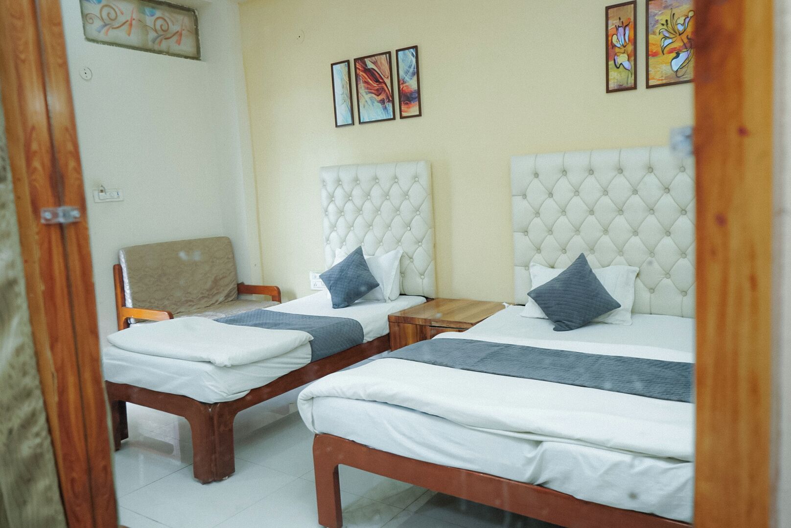 A hotel room with two beds, each with a tufted headboard, wooden frames, and white bedding. There are two bedside tables, one on each bed. The room has a neutral color scheme with beige walls and a wooden door frame.