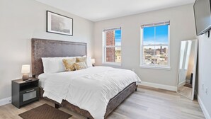 Ein Schlafzimmer mit einem großen Bett, einem Nachttisch, einem gerahmten Bild an der Wand und Blick auf die Stadt durch die Fenster.