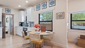 A modern kitchen with white cabinets, a dining table with chairs, and a large window with a view of trees. - Casa Corazón - Walk to UA (Tucson)