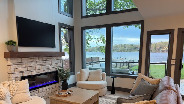 A living room with a stone fireplace, a flat-screen TV, and large windows overlooking a lake. - The Lakehouse on Mona Lake (Norton Shores)