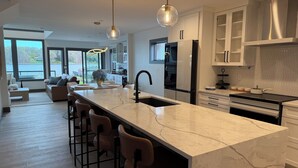 A modern kitchen with a marble island, bar stools, stainless steel appliances, and large windows overlooking a body of water. - The Lakehouse on Mona Lake (Norton Shores)