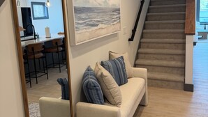 A living room with a sofa, a large mirror, a painting of the ocean, a staircase, and a kitchen in the background. - The Lakehouse on Mona Lake (Norton Shores)