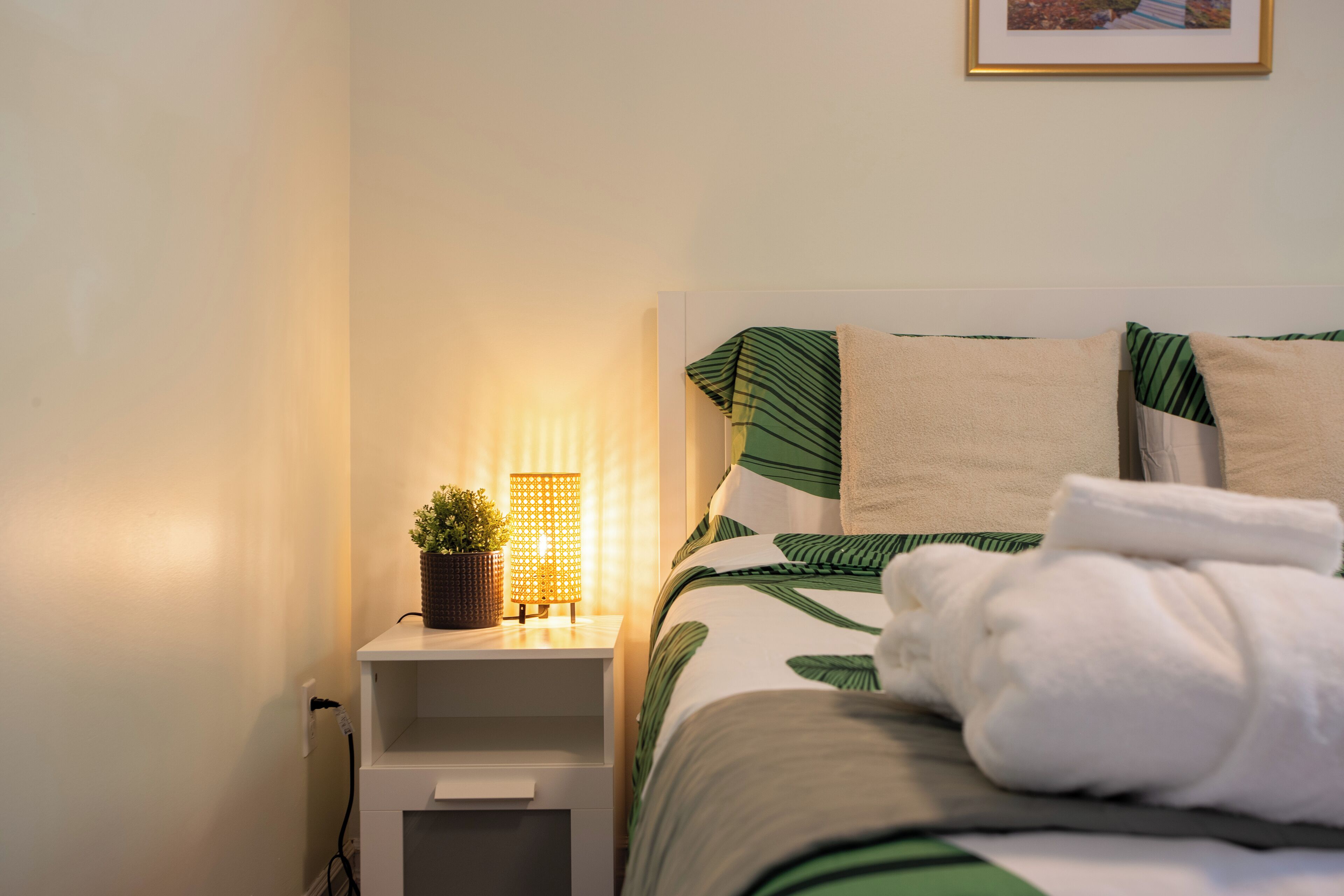A neatly made bed with a green leaf pattern, a bedside lamp, and a small potted plant on the nightstand.
