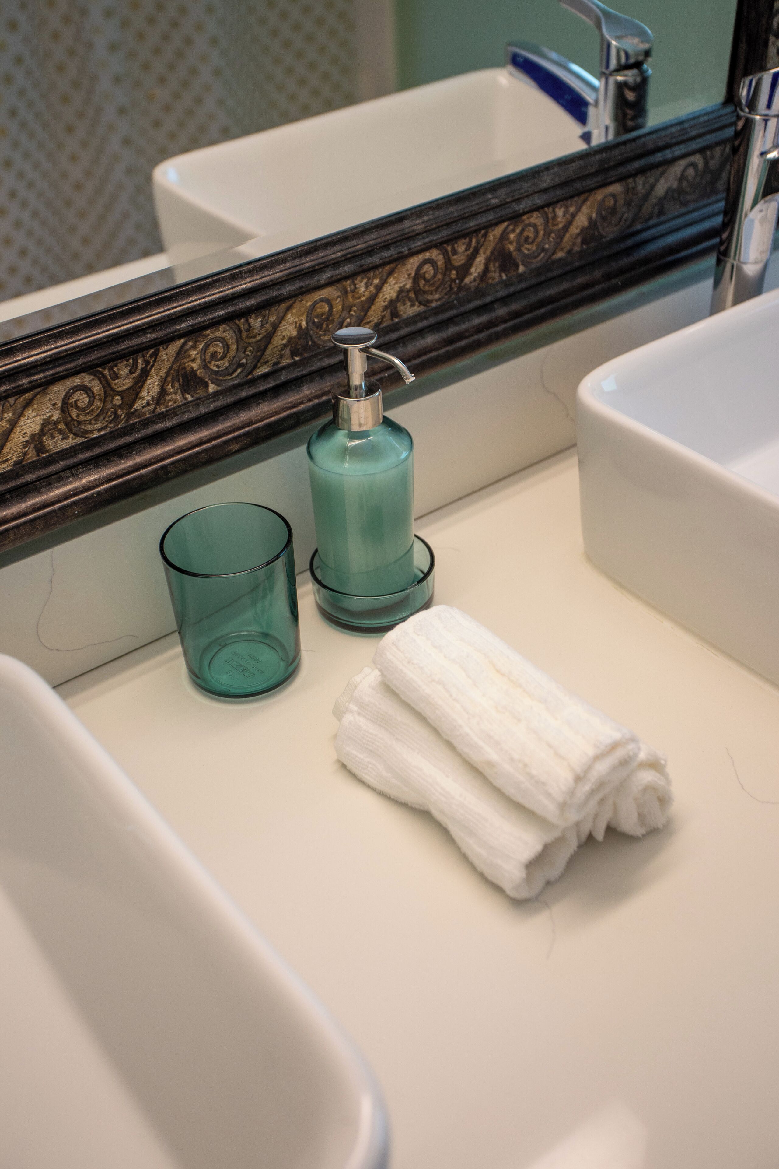 A bathroom with a white sink, a mirror with an ornate frame, a teal soap dispenser, and a white towel.