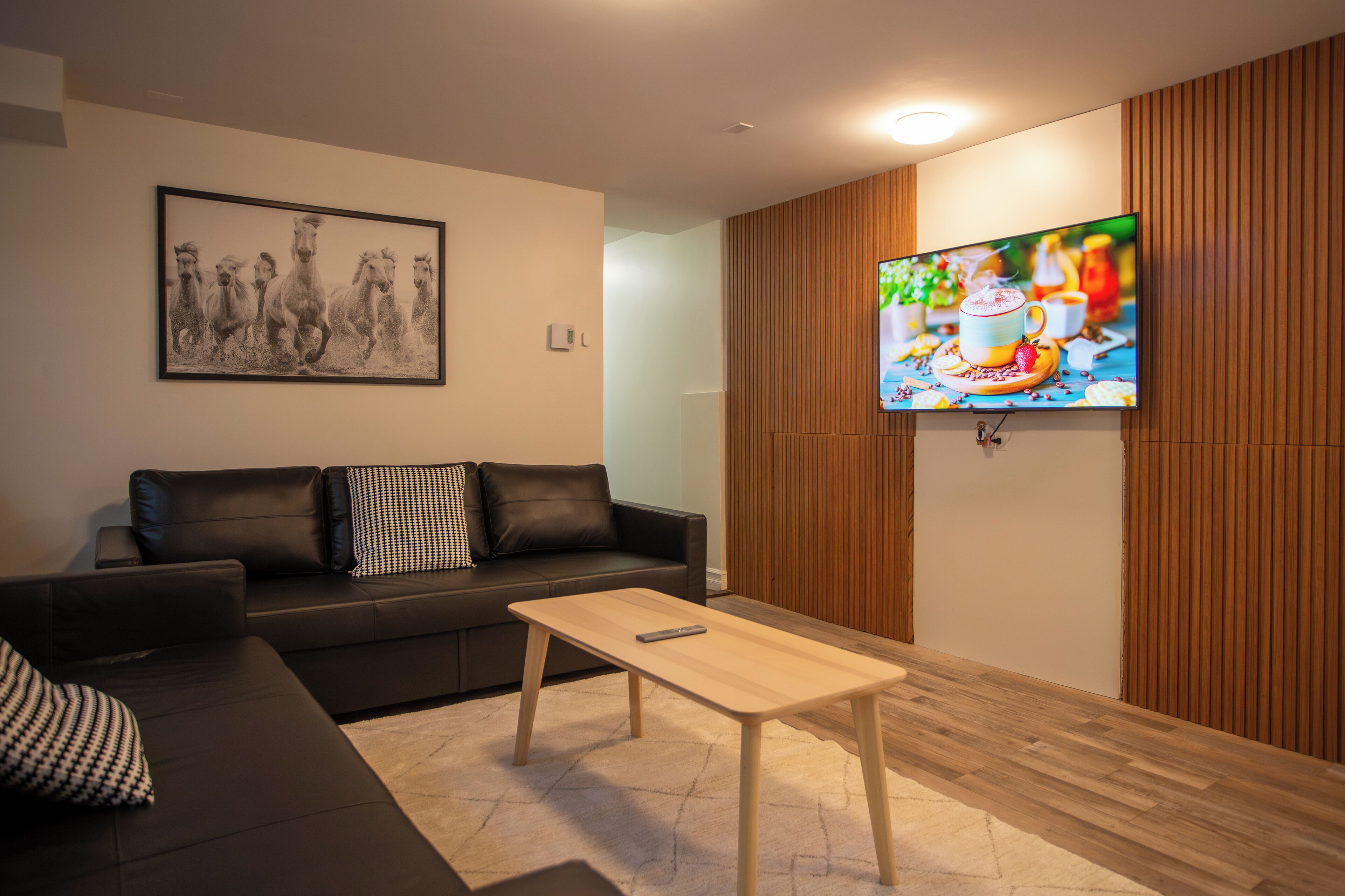 A modern living room with a black leather sofa, a wooden coffee table, and a flat-screen TV mounted on a wooden wall.
