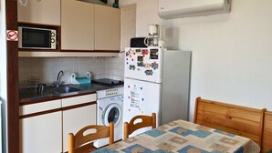 Eine kompakte Küche mit Mikrowelle, Herd, Waschmaschine und Kühlschrank, ein Esstisch mit Stühlen sowie ein hölzernes Bettgestell.