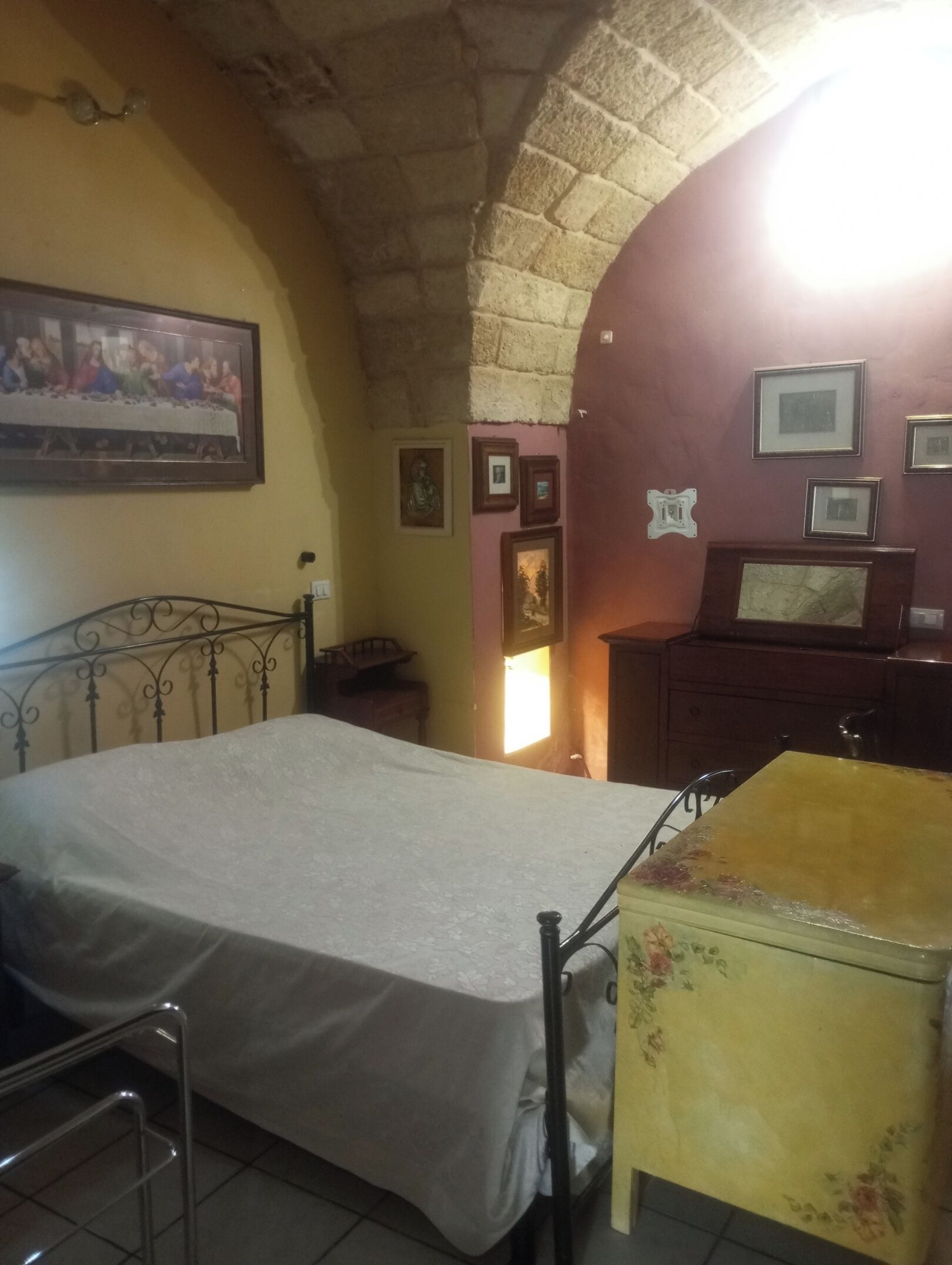 Ein Schlafzimmer mit Bett, kleinem Tisch und Kommode. Ein Gemälde hängt an der Wand, und durch das Fenster scheint Sonnenlicht.