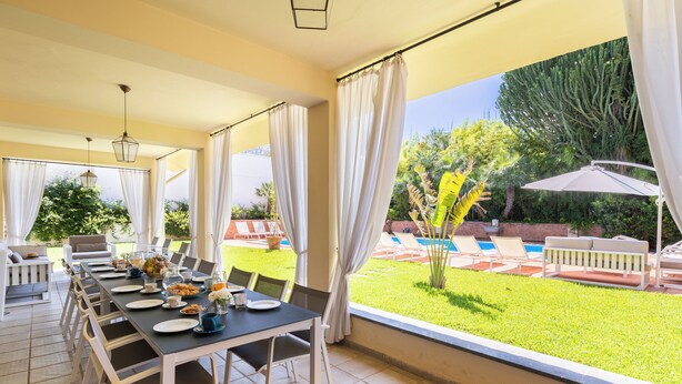 Uma sala de jantar com mesa posta para quatro pessoas, janelas amplas e vista para uma piscina e jardim.