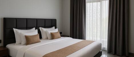 A modern hotel room with a large bed, dark headboard, and a view of the city through sheer curtains.