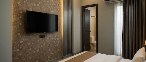 A hotel room with a flat-screen TV mounted on a floral-patterned wall, a bed with a beige and brown bedspread, a bathroom with a toilet and shower, and a window with dark curtains.