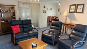 Un salon comprenant un canapé, deux fauteuils, une table basse, une bibliothèque et, au fond, un coin repas.