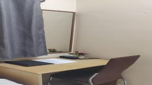 Une pièce de taille modeste comprenant un lit, un bureau avec une chaise, un miroir et une petite table sur laquelle est posé un vase de fleurs.