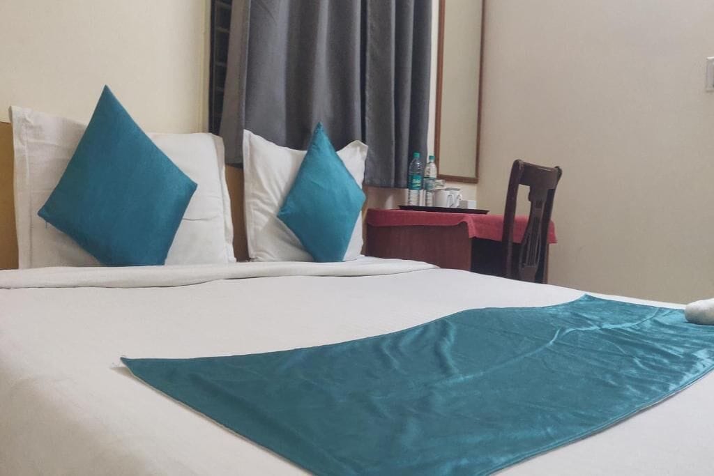 A hotel room with a bed, two pillows, a table with a red tablecloth, and a chair.