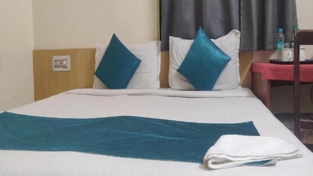 Une chambre d’hôtel comprenant un lit, deux oreillers, une couverture, une serviette pliée, une table de chevet, une bouteille d’eau et une machine à café.