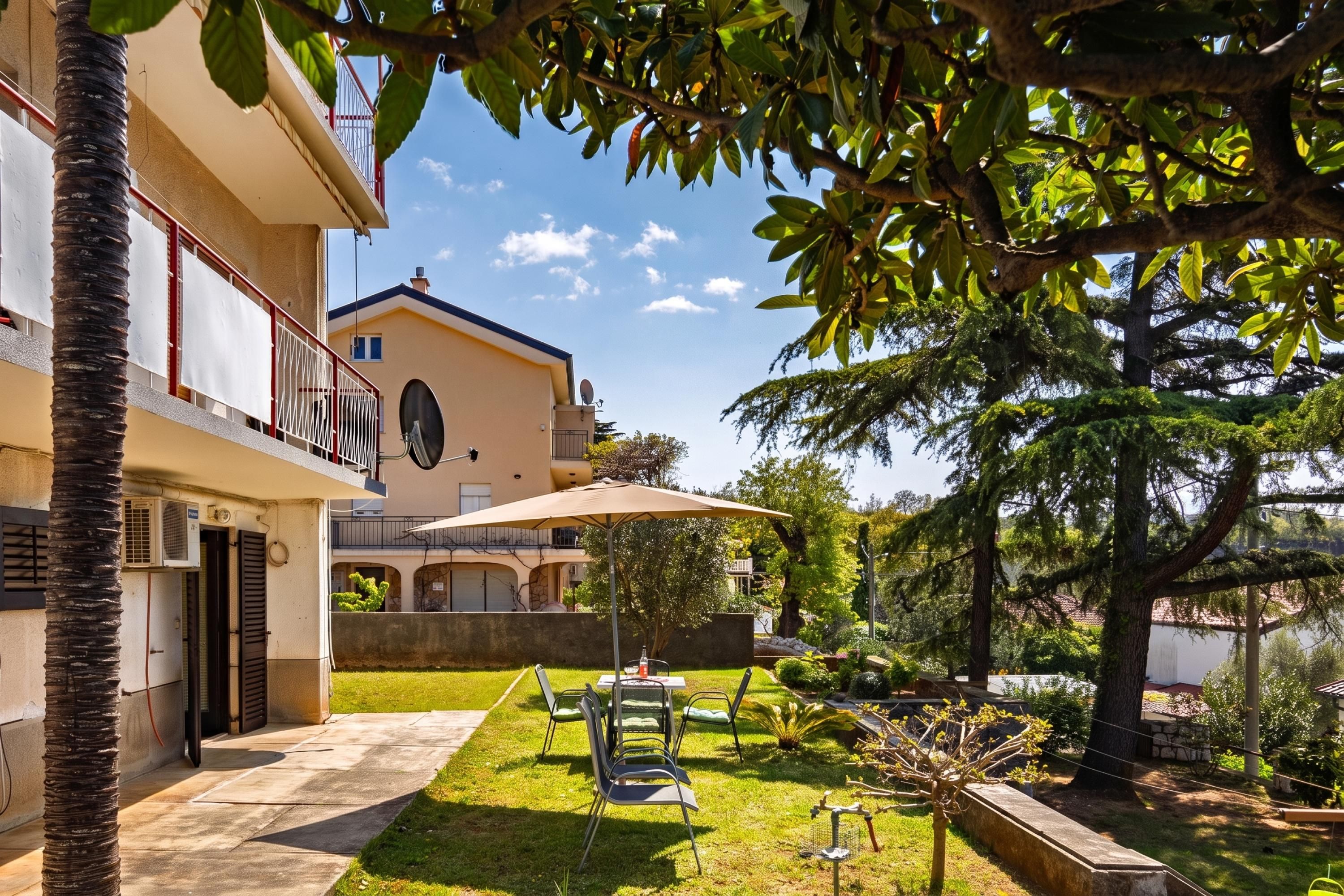 Un bâtiment doté d’un balcon, d’un patio avec une table et des chaises, et d’un jardin avec des arbres et une pelouse.