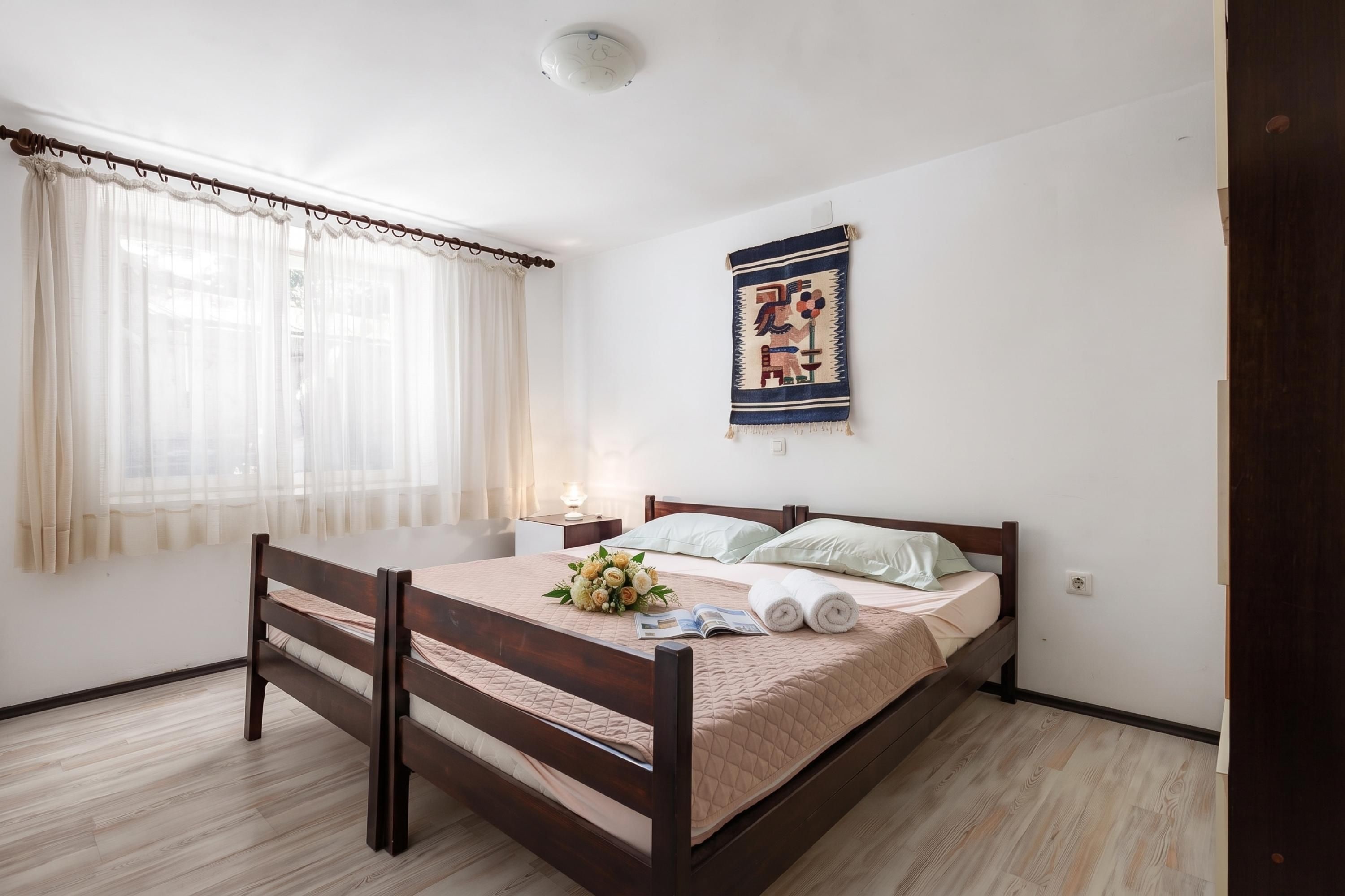 A bedroom with a wooden bed, a bedside table with a lamp, a window with sheer curtains, and a wall hanging.